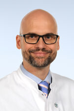Porträtfoto eines Mannes mit Glatze und Bart. Er trägt eine Brille mit dunklem Brillengestell, ein hellblaues Hemd und eine gestreifte Krawatte. Darüber trägt er einen weißen Arztkittel und lächelt freundlich in die Kamera.