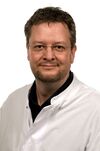 Porträtfoto eines Mannes mit dunklen Haaren. Unter seinem weißen Arztkittel trägt er ein dunkles Oberteil und lächelt freundlich in die Kamera.