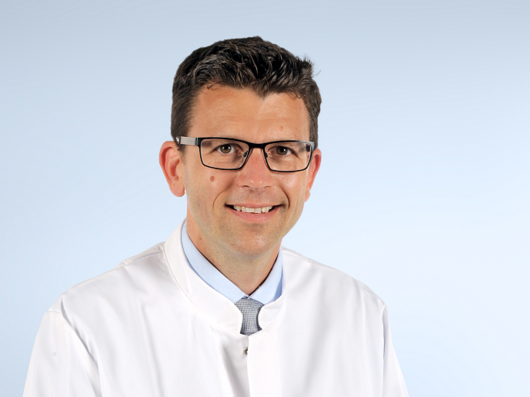 Porträtfoto eines Mannes mit kurzen, dunklen Haaren und einer Brille mit dunklem Brillengestell. Er trägt ein hellblaues Hemd und einen weißen Arztkittel. Er lächelt freundlich in die Kamera.