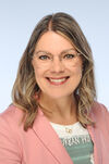 Porträtfoto einer Frau mit schulterlangen braun-blonden Haaren, die sich leicht wellen. Sie trägt eine Brille, ein weißes T-Shirt mit grünem Print und einen rosefarbenen Blazer. Sie lächelt freundlich in die Kamera.